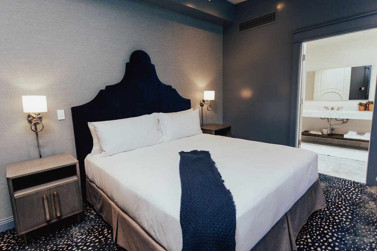 A hotel room with a large bed, bedside tables, a nightstand, a mirror, and a bathroom visible through an open door.