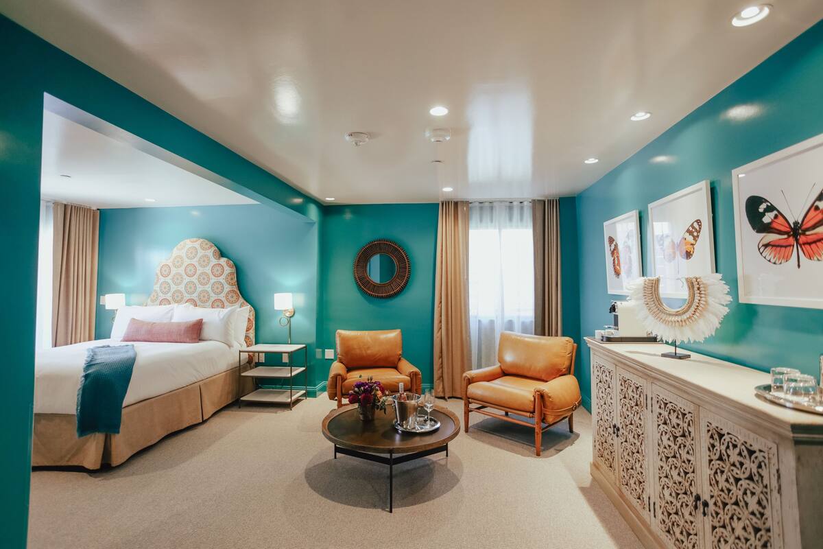 A hotel room with teal walls, a bed with a patterned headboard, two leather armchairs, a coffee table with a tray, and a decorative mirror.