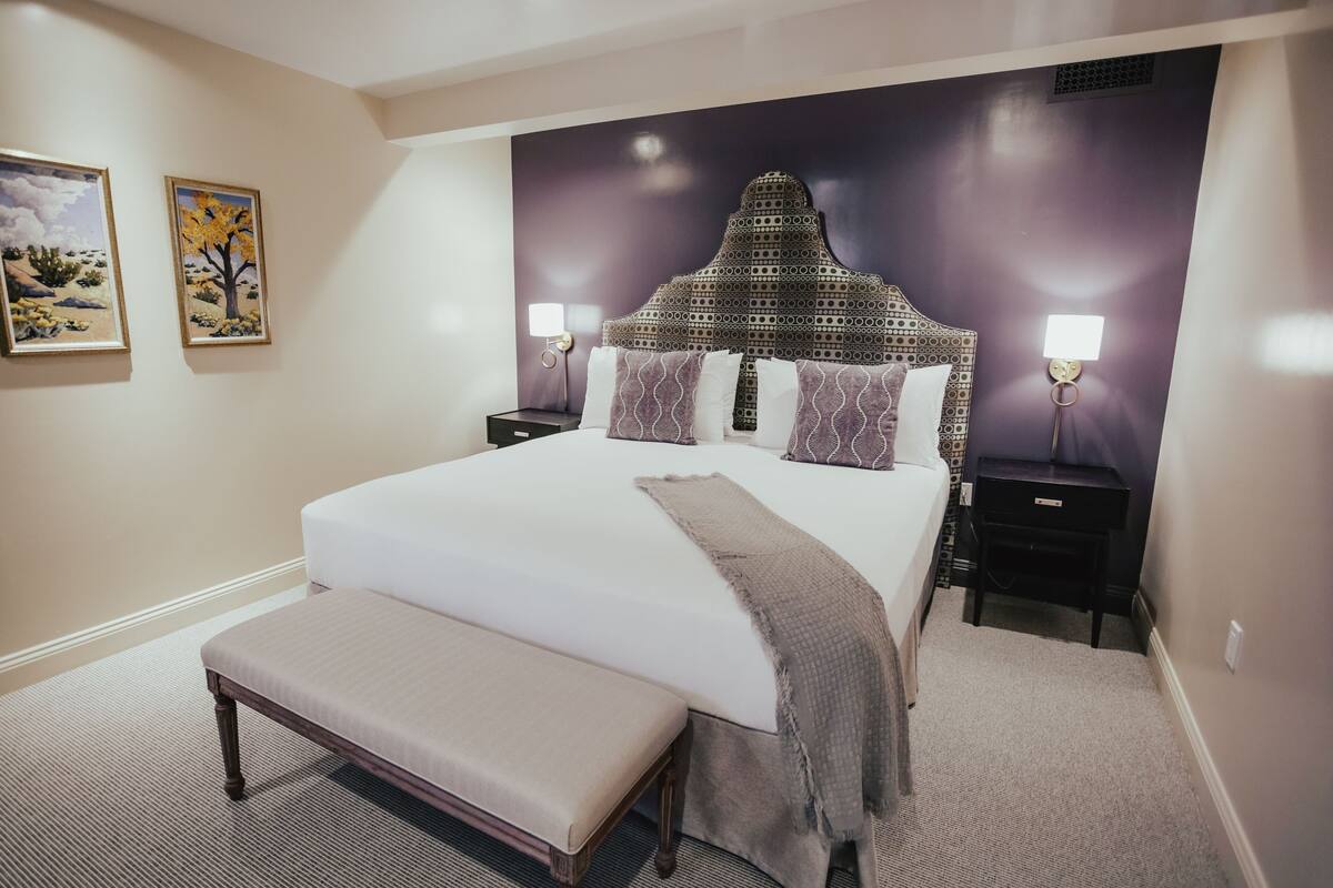 A bedroom with a large bed, a bench, two nightstands, and wall art.