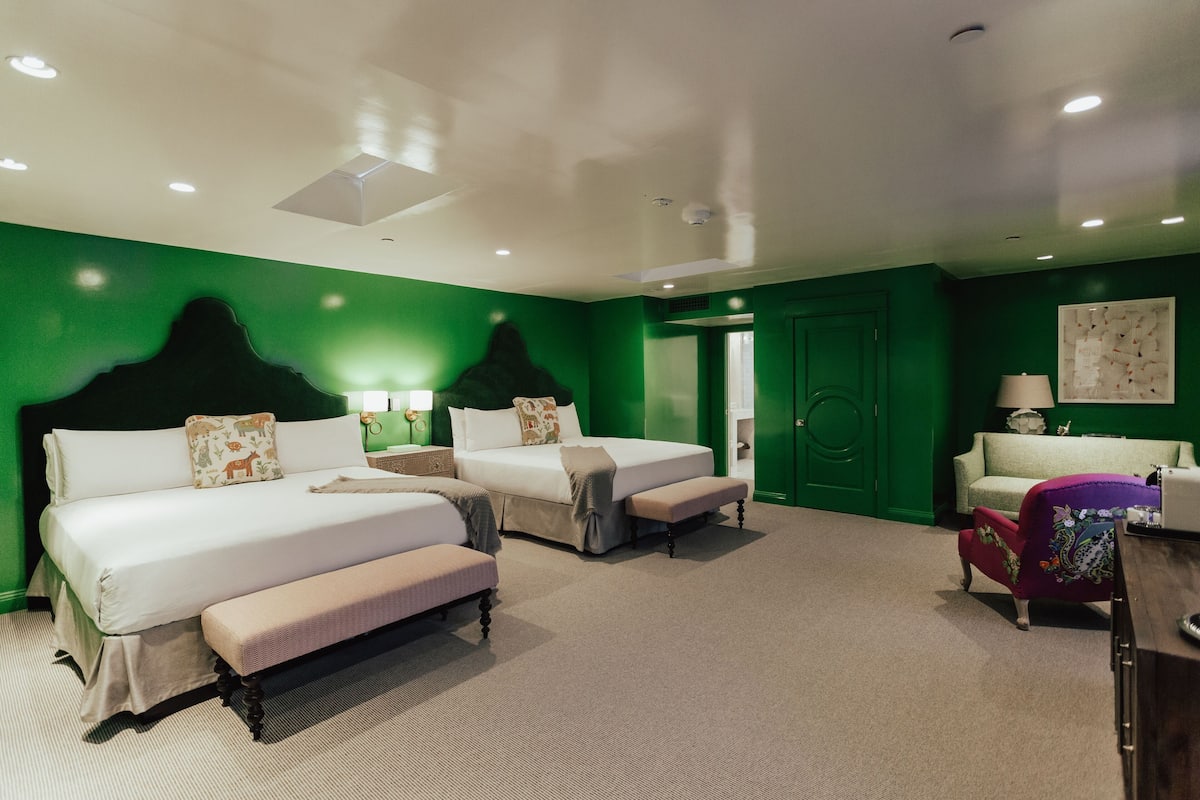 A hotel room with two beds, a bench, a chair, and a green wall.