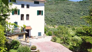 A white building with green shutters, a paved courtyard, and a mountainous backdrop. - Nice apartment for 3 people with pool, WIFI, patio, pets allowed and panoramic view (Lecchiore)