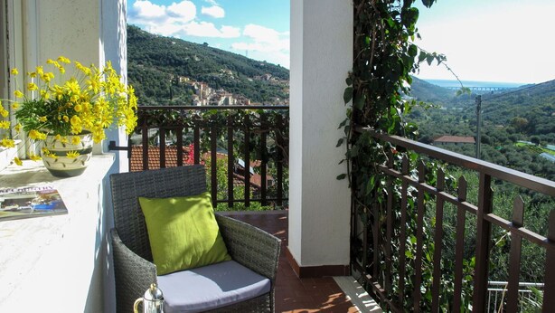 A balcony with a wicker chair, a table with a tray set, and a potted plant with yellow flowers. - Nice apartment for 6 guests with WIFI, pool, TV and patio (Dolcedo)