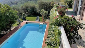 A rectangular swimming pool with a wooden deck, surrounded by lush greenery and a view of a hilly landscape. - Nice apartment for 6 guests with WIFI, pool, TV and patio (Dolcedo)