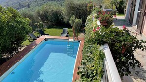 A rectangular swimming pool with a wooden deck, surrounded by lush greenery and a view of a hilly landscape. - Cosy apartment for 10 guests with private pool, WIFI, TV and patio (Dolcedo)