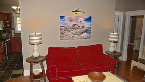 Ein Wohnzimmer mit einem roten Sofa, zwei Beistelltischen mit Lampen, einem Holztisch mit einer Schale und einem Gemälde eines Leuchtturms an der Wand.