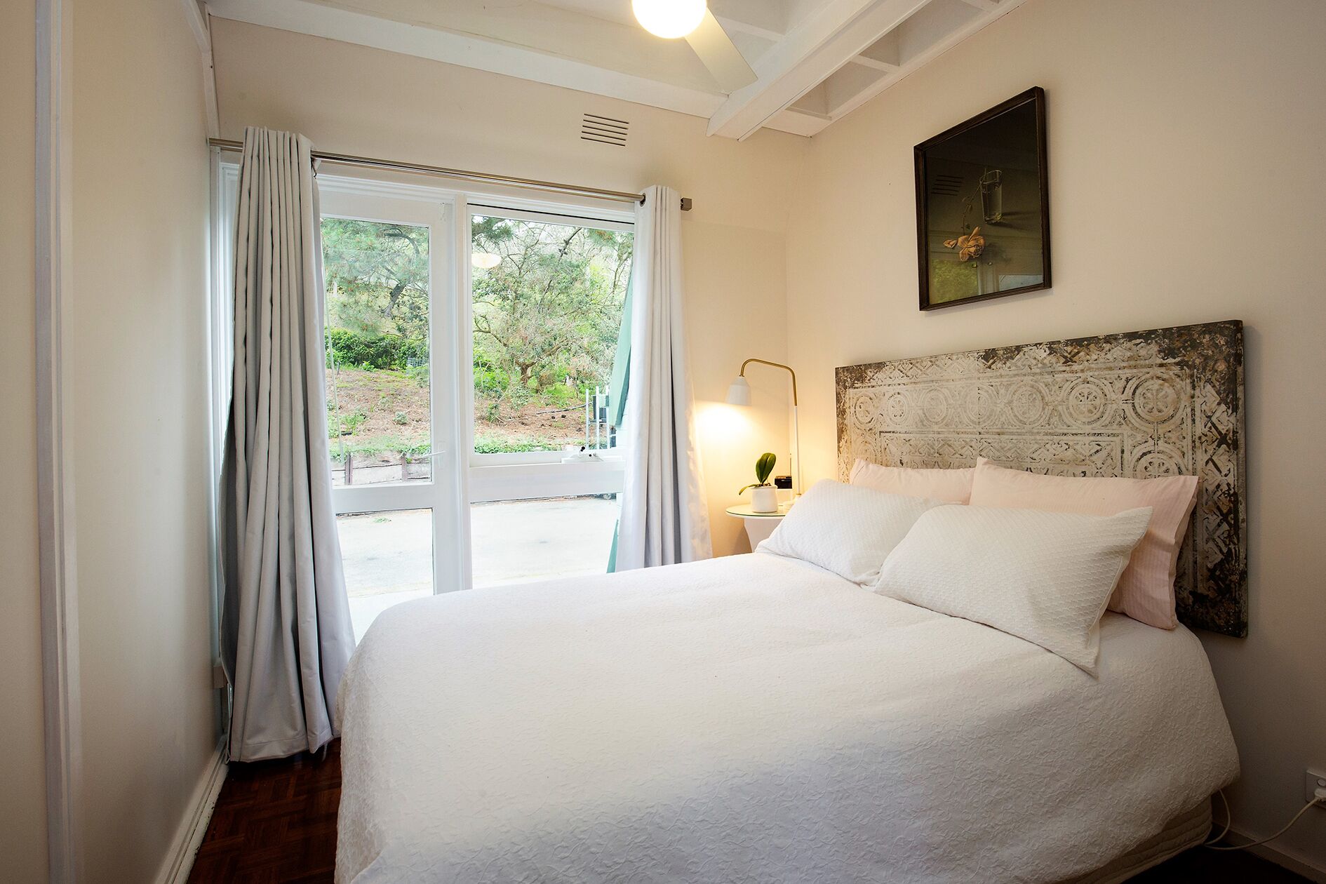 A bedroom with a large bed, a headboard with a textured design, a bedside lamp, and a view through a glass door to an outdoor area.