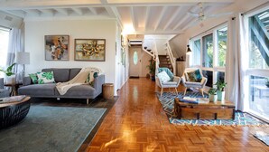 Amplia sala de estar con piso de madera en espiga, un sofá gris y una pequeña mesa de centro. Se ve una escalera que conduce a un nivel superior, y ventanas amplias a la derecha.