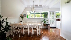 Un comedor con una mesa de madera y sillas blancas, una lámpara colgante y una ventana con vistas a follaje.