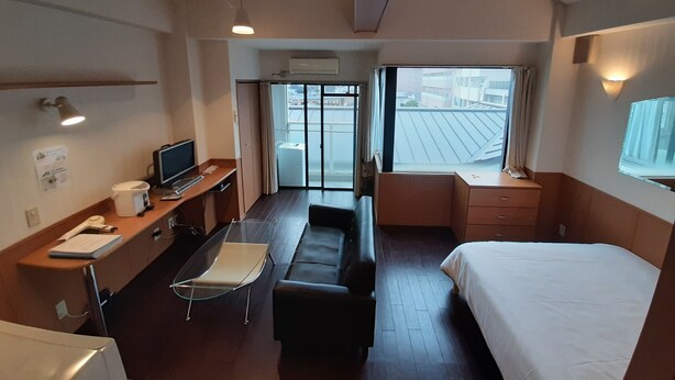 Ein Hotelzimmer mit einem Bett, einem Schreibtisch mit Computer, einem Sessel, einer Couch und einem Fenster mit Blick auf Gebäude.