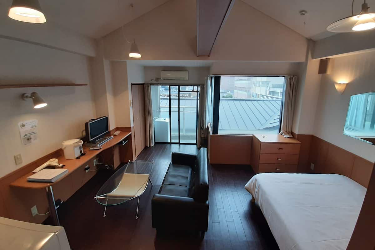 Quarto de hotel com cama, escrivaninha com computador, cadeira, sofá e janela com vista para edifícios.