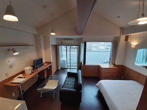 A hotel room with a bed, a desk with a computer, a chair, a sofa, and a window with a view of buildings.