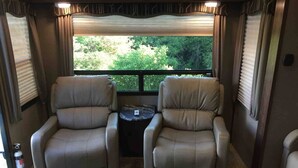 A cozy RV interior with two reclining chairs and a small table, a window with curtains, and a fire extinguisher on the wall. - Camper at the Sequoia Forest Retreat (Badger)