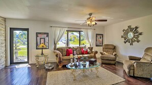 A spacious living room with a glass coffee table, a sofa, and two armchairs. There is a ceiling fan, a large mirror, and a door leading to a balcony. - Pet-Friendly Oasis w/ Deck & Fire Pit in Bokeelia! (Bokeelia)