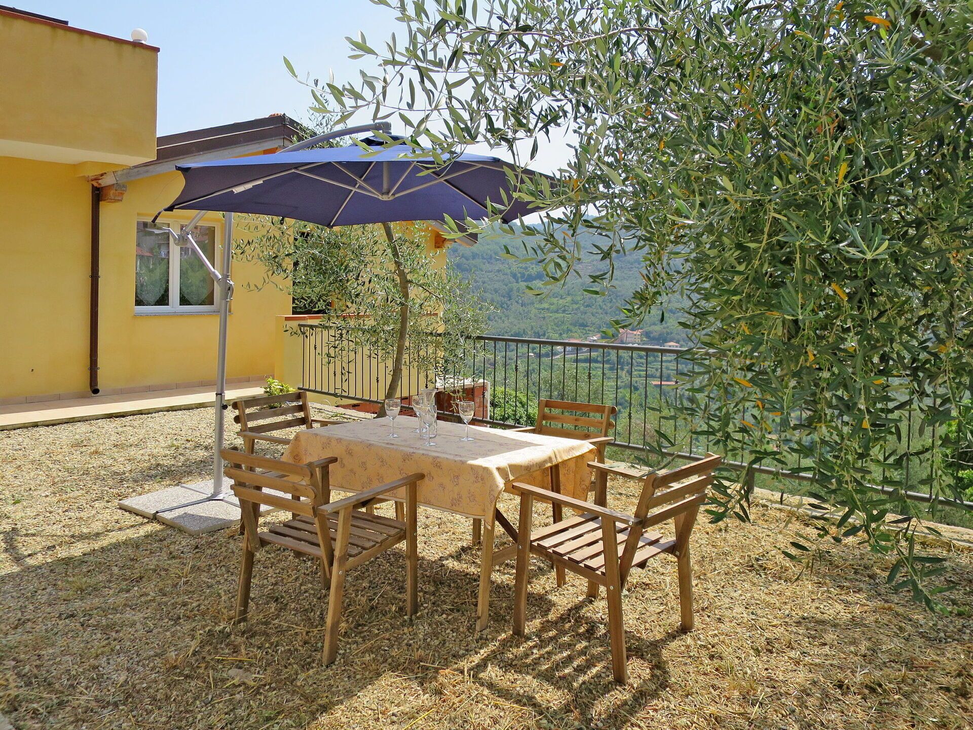 Un patio exterior con mesa y sillas, una sombrilla azul y vistas a una colina.