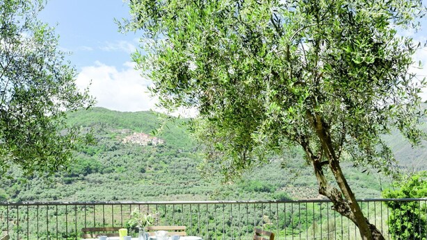 Uma área de refeições ao ar livre com uma mesa posta para quatro pessoas, cercada por cadeiras de madeira, com vista para um vale verdejante e uma aldeia ao longe.