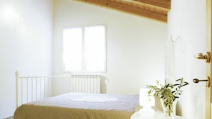 Um quarto com teto de madeira, cama branca com colcha bege, janela com persianas e radiador branco.