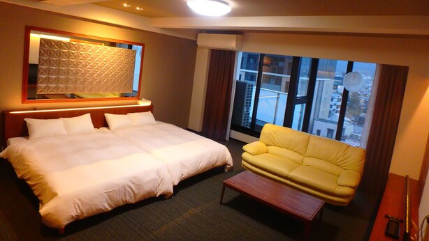 Habitación de hotel con una cama grande, un sofá amarillo, una pequeña mesa de centro de madera y un ventanal con vistas a la ciudad.