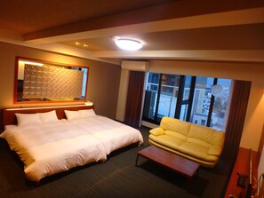A hotel room with a large bed, a yellow sofa, a small wooden coffee table, and a large window offering a city view.