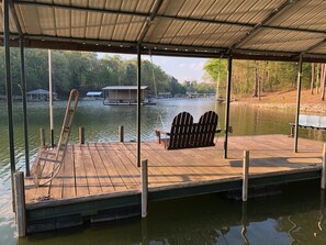 Un muelle de madera con una silla hamaca y un banco, con vista a un lago tranquilo, una casa anclada y árboles al fondo.