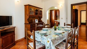 A dining area with a table set for four, wooden chairs, a television, and a cabinet with glass doors. - Beautiful apartment for 6 guests with WIFI, pool, TV, terrace and pets allowed (Diano Castello)