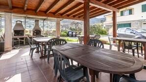 Une terrasse extérieure avec des tables et des chaises en bois, un four en pierre et une vue sur une montagne et des bâtiments en arrière-plan.