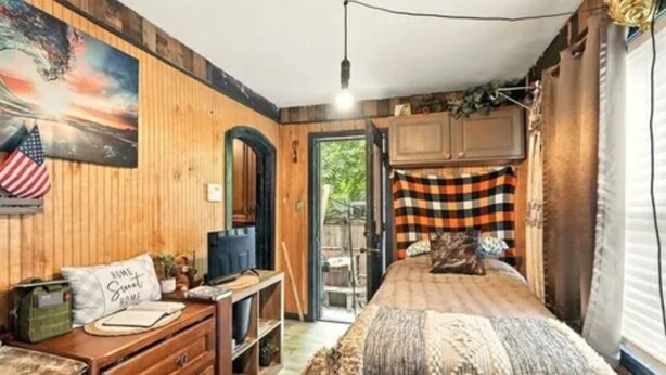 Une chambre avec un mur en bois, un lit recouvert d’une couverture à carreaux, une commode, un miroir et un tableau.