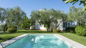 Une piscine entourée d’une végétation luxuriante et d’arbres, avec un bâtiment blanc en arrière-plan.