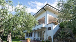 Une maison à deux étages dotée d’un balcon, entourée d’une végétation luxuriante et d’un mur de pierre.
