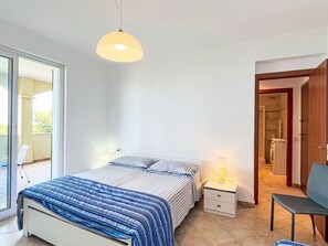 Ein Schlafzimmer mit Bett, Nachttischen, einer Lampe, einem Sessel und Blick auf einen Balkon sowie einen Wäschebereich.
