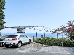 Ein weißes Auto parkt auf einer Einfahrt mit gepflastertem Bereich, einem Hinweisschild und Blick auf das Meer im Hintergrund.