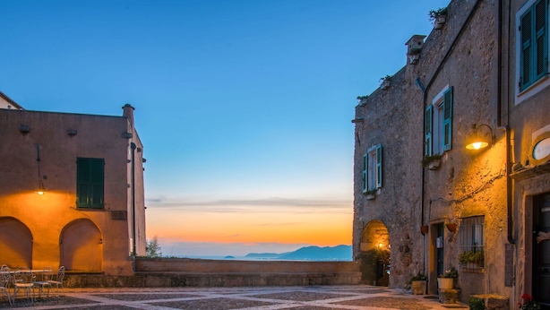 A cobblestone courtyard with buildings on either side, featuring arched doorways and a view of distant mountains at sunset. - Cosy apartment for 5 guests with WIFI, TV, terrace and pets allowed (Pietra Ligure)