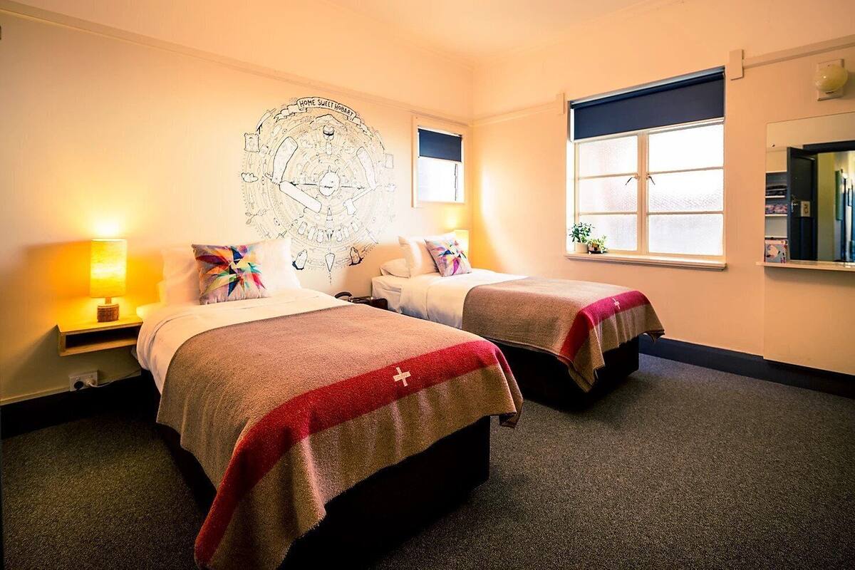 A hotel room with two beds, a large wall tapestry, bedside lamps, and a window with blinds.