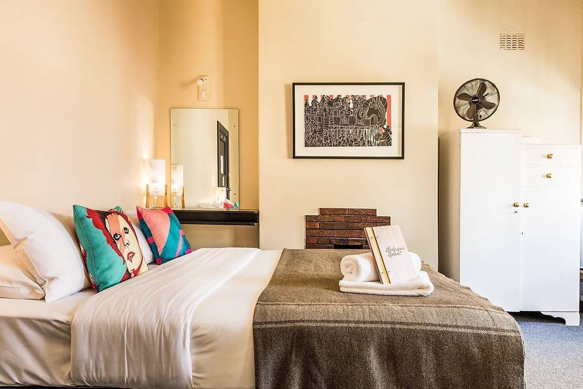 A hotel room with a bed, pillows, a bedside table with a lamp, a mirror, a framed artwork, a fan, and a wardrobe.
