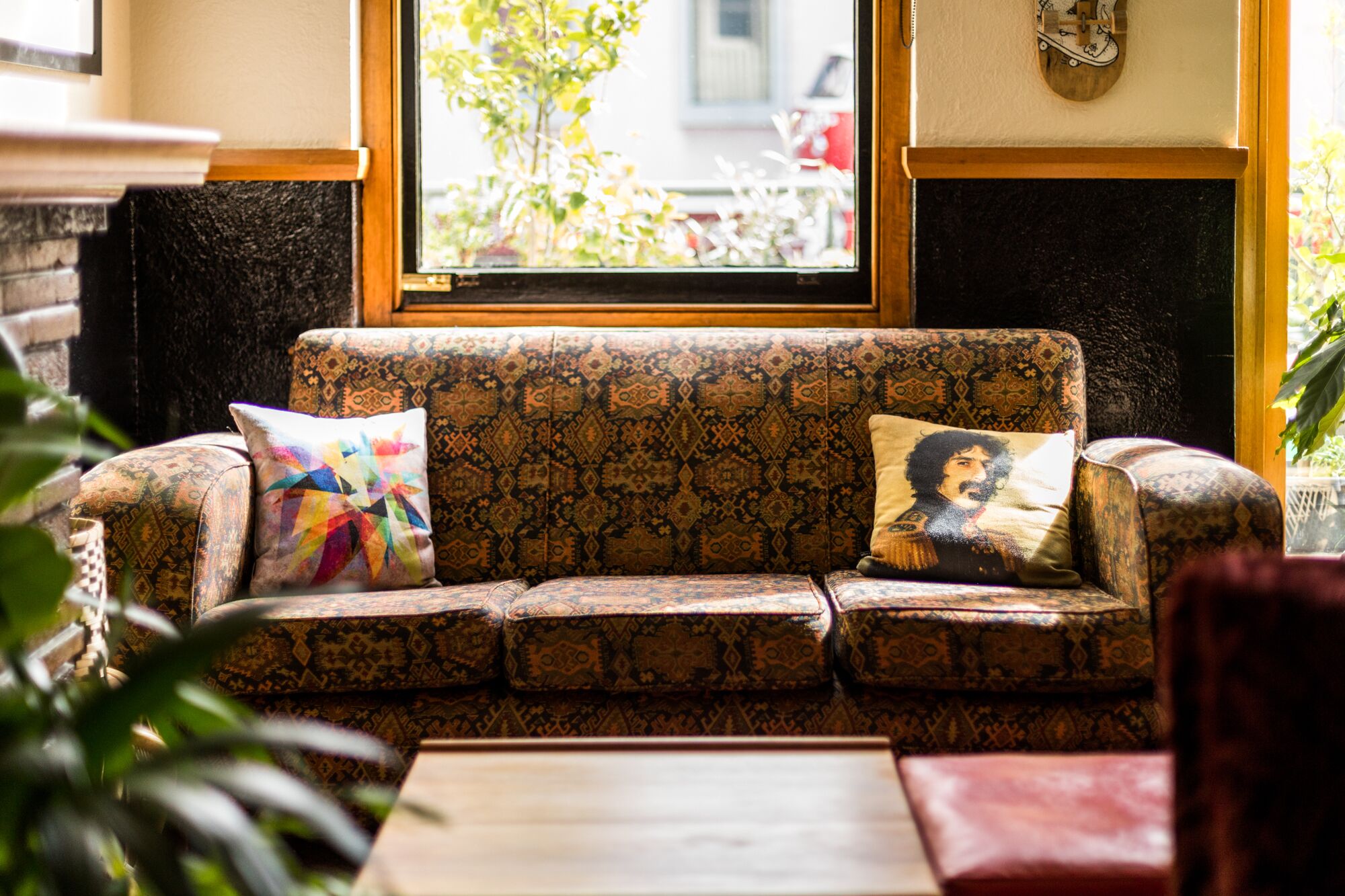 A patterned sofa with two cushions, a window with a view of greenery, and a wooden table in a room with a brick wall and a black fireplace.