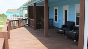 A covered deck with wooden flooring, a seating area with two chairs and a table, and a view of a blue house in the background. - Shore Fun! 3/2 Beachhouse, Holiday Beach Subdivision, Close to Everything! (Port Bolivar)