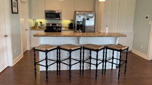 A kitchen with white cabinets, a stainless steel refrigerator, a microwave, and a stove. There are four wooden stools with cushioned seats. - Shore Fun! 3/2 Beachhouse, Holiday Beach Subdivision, Close to Everything! (Port Bolivar)