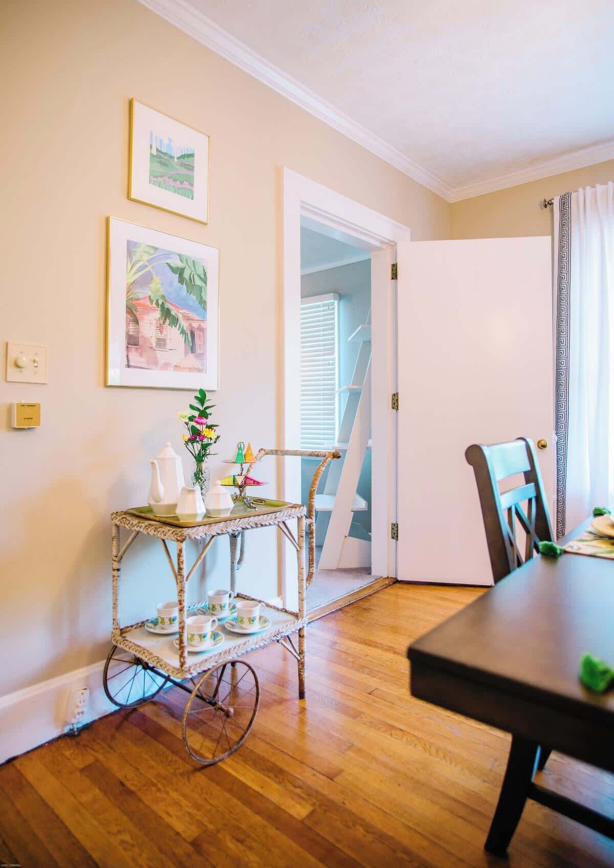 A dining area with a wooden table and chairs, a wicker cart with tea set, and two framed pictures on the wall.