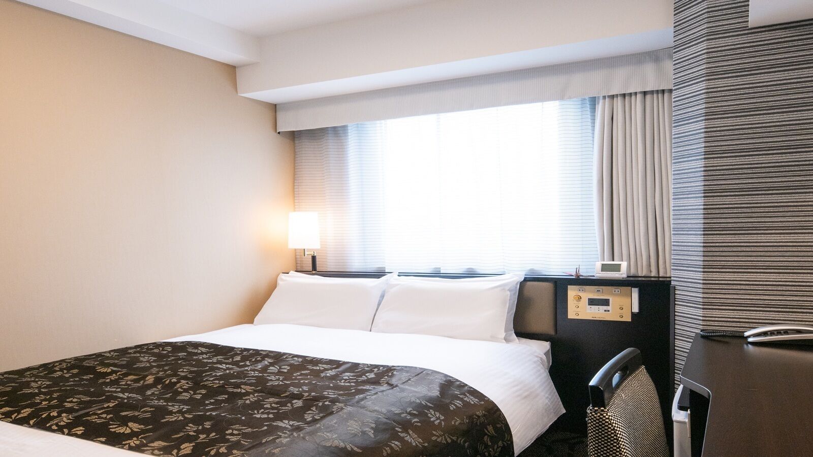 A hotel room with a bed, a desk, a chair, a window with blinds, and a control panel.