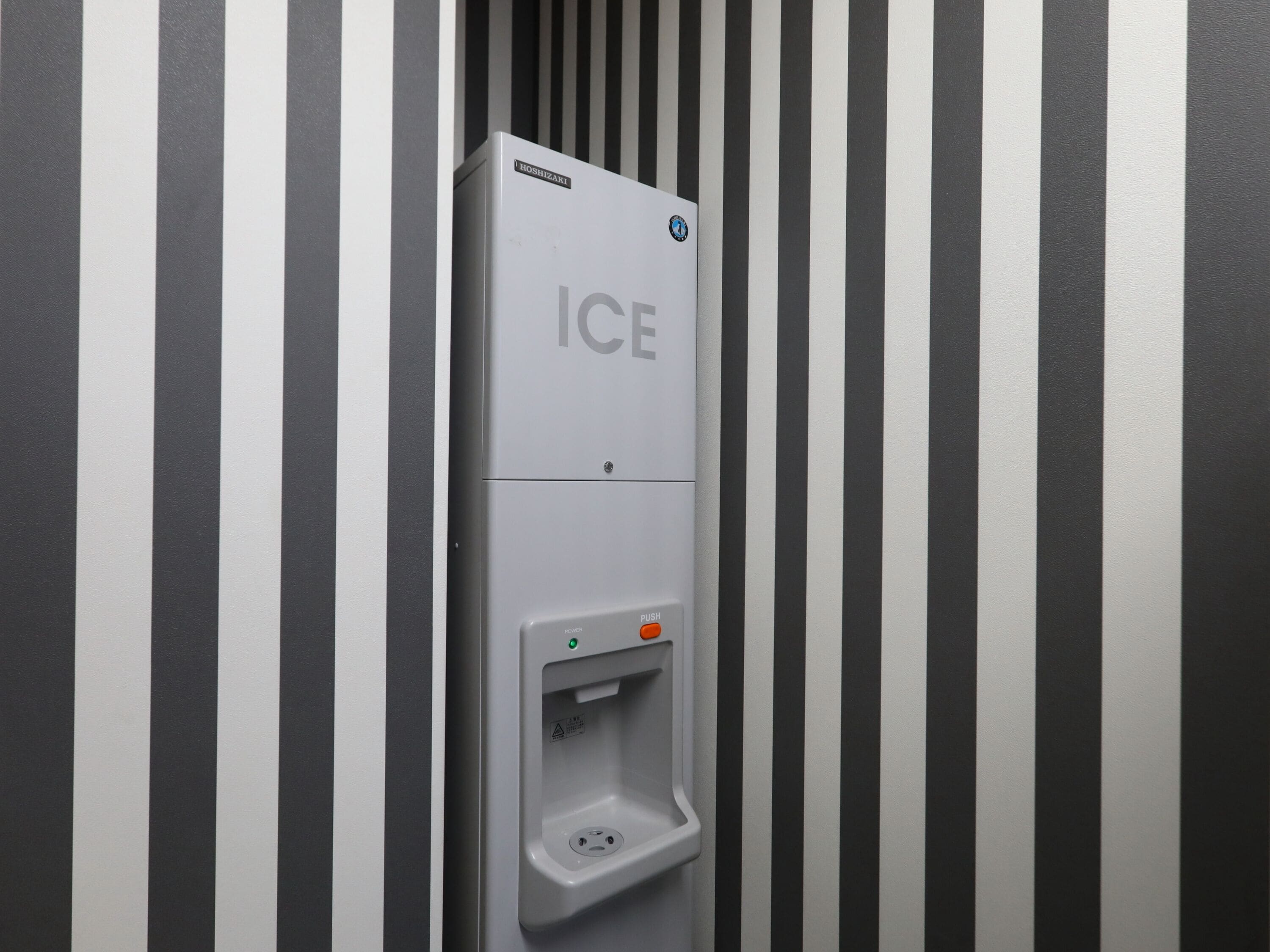 Máquina expendedora con etiqueta que dice "ICE", un botón de encendido y un botón pulsador, frente a una pared a rayas.