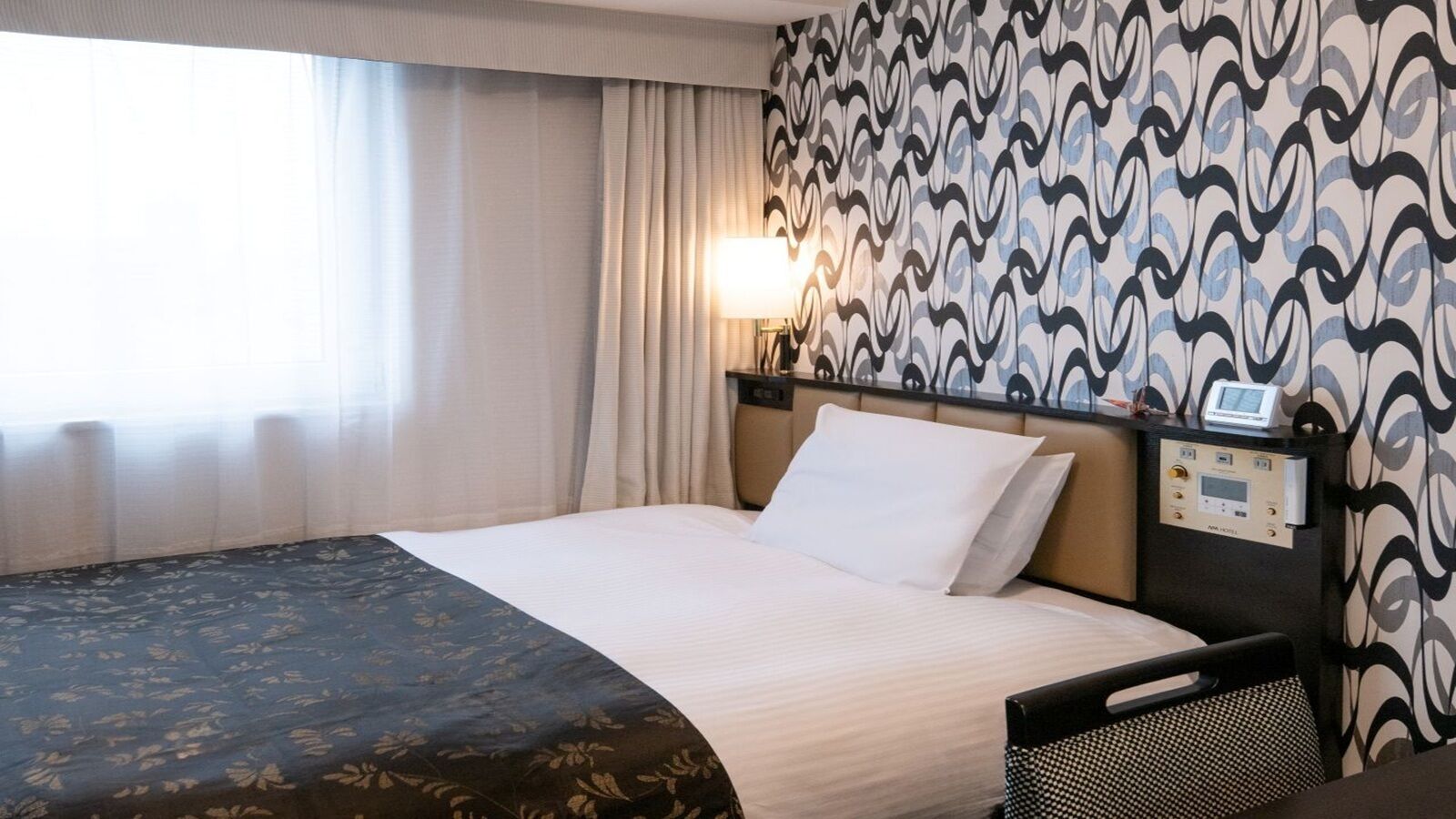 A hotel room with a bed, a bedside table with a lamp, a digital clock, and patterned wallpaper.