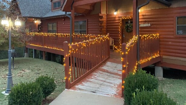 A log cabin with a porch decorated with string lights, a wooden fence, and a small pathway leading to the entrance. - Cozy Cabin 2 - A Great Holiday Family Place - Come Play and Stay! (Branson)