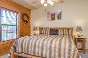 Une chambre à coucher avec un lit en bois, un ventilateur de plafond, une fenêtre avec des stores, une couronne décorative accrochée au mur, un tableau représentant des ours et une table de chevet avec une lampe.