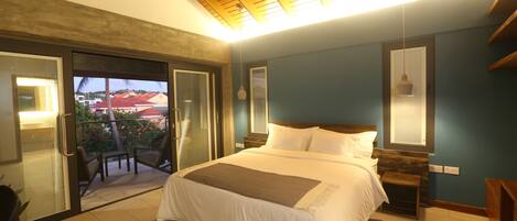 A bedroom with a large bed, a wooden ceiling, a view through a glass door, and a small table with a lamp.