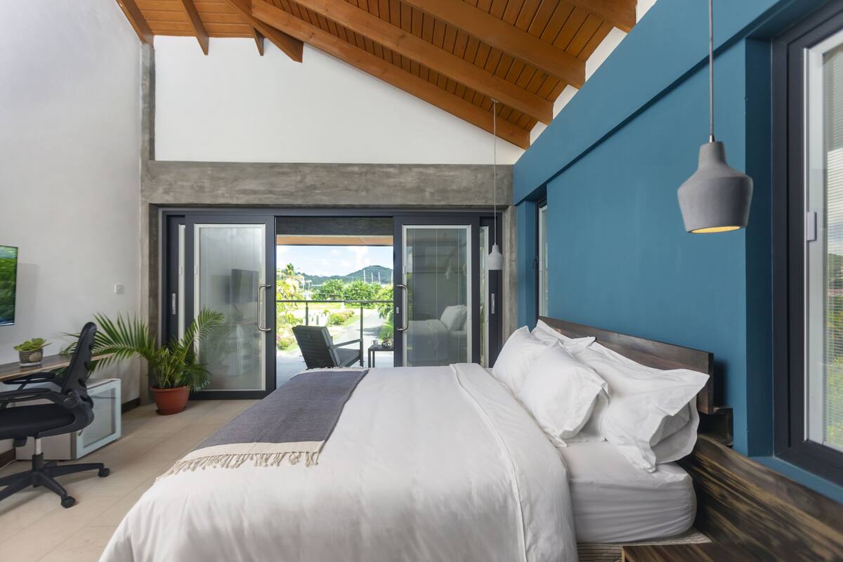 A modern bedroom with a large bed, a desk, and a view of the outdoors.