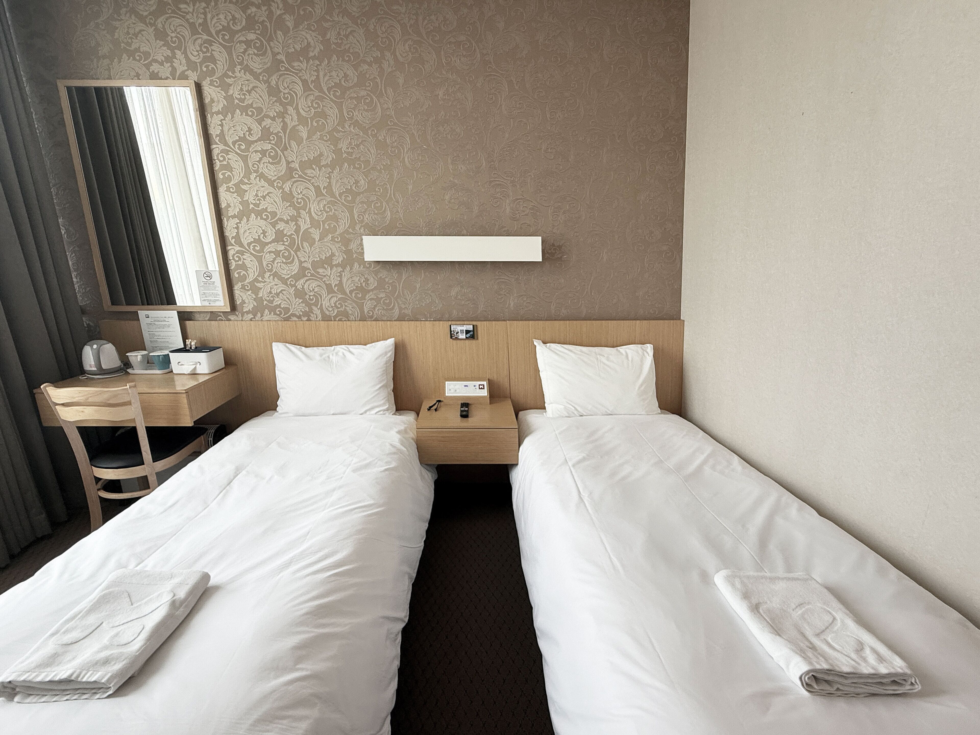 A hotel room with two single beds, a desk with a chair, a nightstand, a mirror, and a wall-mounted control panel.