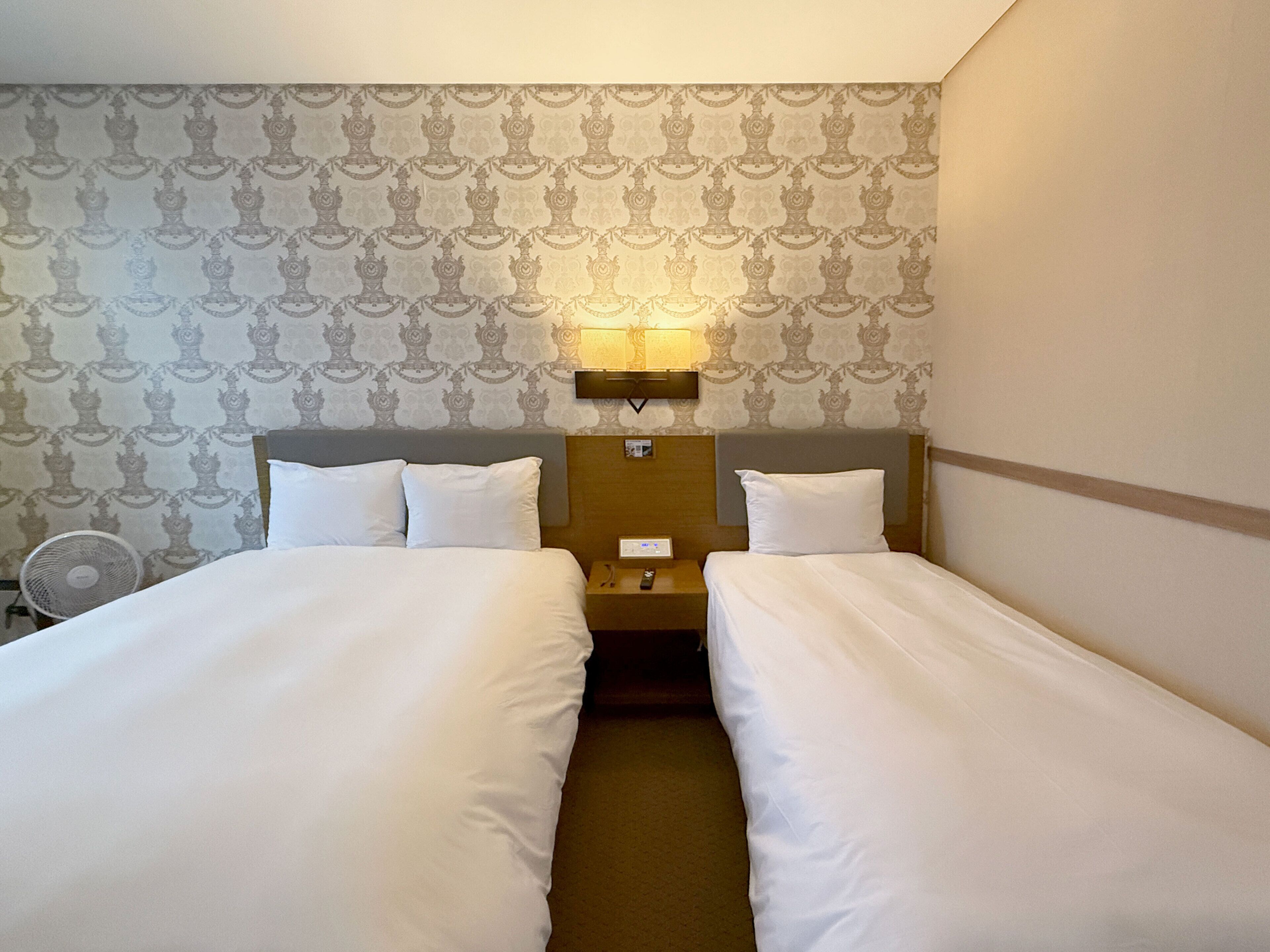 A hotel room with two single beds, a bedside table with a lamp, and a wall-mounted fan.