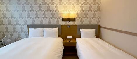 A hotel room with two single beds, a bedside table with a lamp, and a wall-mounted fan.