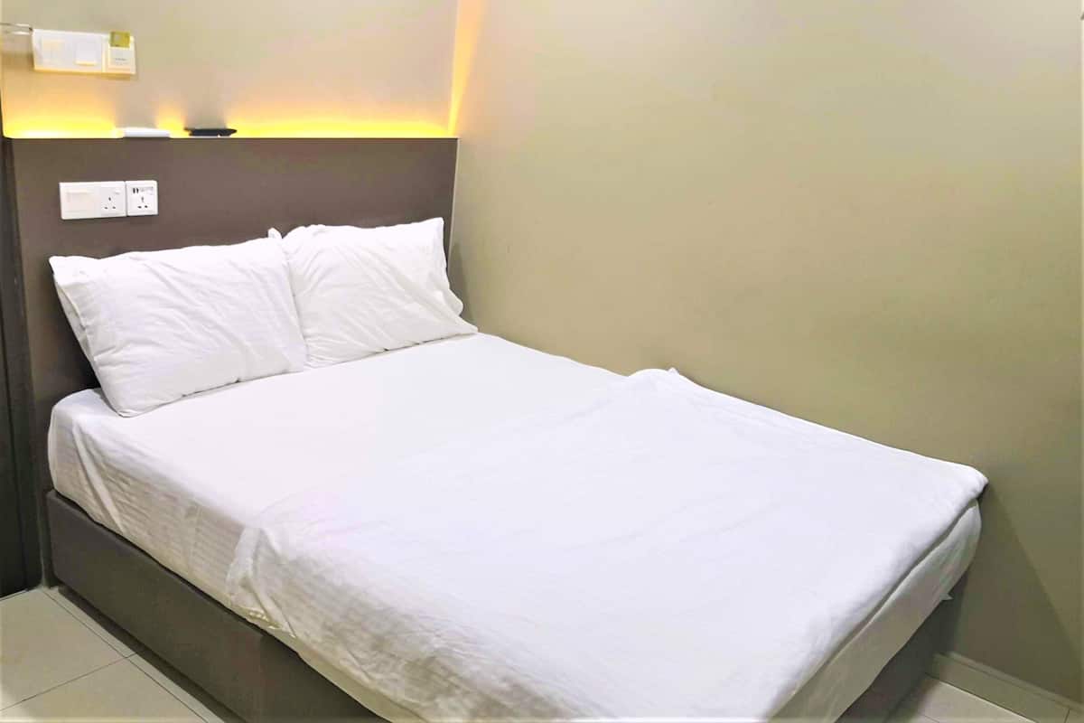 Un lit simple avec une literie blanche, dans une pièce dont le mur est uni et où se trouve un interrupteur juste au-dessus.