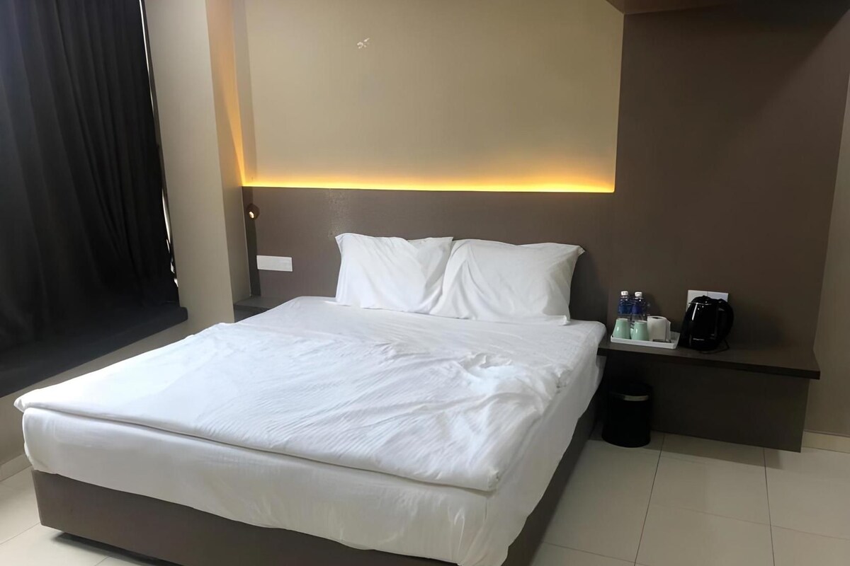 Ein ordentlich bezogenes Bett mit weißen Leinen, ein Nachttisch mit Kaffeemaschine und Pflegeprodukten sowie eine an der Wand befestigte Leuchte.