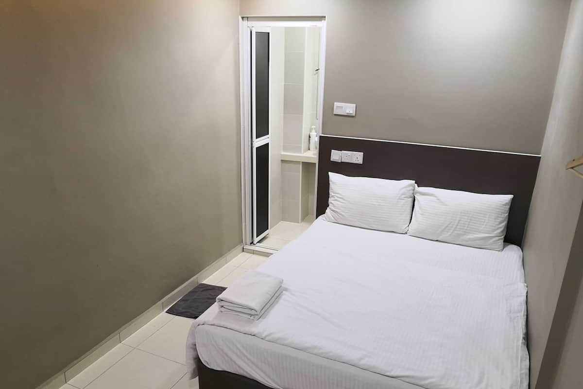 Un lit bien fait, avec du linge de lit blanc, dans une petite chambre moderne dotée d’une tête de lit sombre et d’une porte donnant accès à une salle de bain.